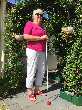  [image description] A lovely photograph of Carol standing, smiling, with her long white cane fully extended in a vertical placement in front of her body. She is wearing white summer pants and a bright pink t-shirt and is surrounded by a lovely green garden setting