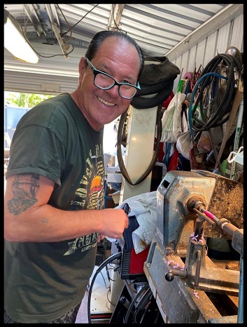 [image description] A terrific half side on body profile of Tony standing at his workshop bench creating a pen and smiling towards the camera. He is wearing a pair of glasses and equipment can be seen positioned on his workshop bench
