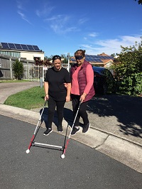 Two Support Workers on Professional Development training, one guiding the other under blindfold, with two long canes joined by a horizontal bar, both stepping off a kerb edge