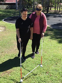 Two Support Workers on Professional Development training, one guiding the other under blindfold, with two long canes joined by a horizontal bar, both walking on grass in a park