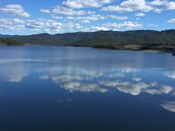 a beautiful photo of Hinze dam with blue sky, clouds and distant mountain range reflecting on water's surface