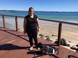 Hailey standing on boardwalk with Seeing Eye Dog Orella lying down and sand and ocean in background