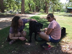 Jessie and Tarhnee sitting down on grass in a shady parkland, with Jessie patting dog guide who is standing