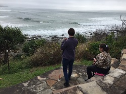 Image shows Gabbi standing from behind and Hailey sitting from behind facing ocean expanse ahead of them