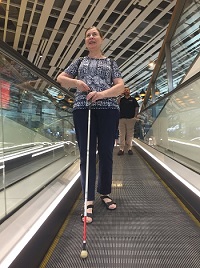 Anne-Marie seen with long cane ahead and standing whilst descending a travelator