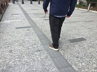 Photo taken from behind and to the left side of a male who is seen walking along a set of TGSIs in King George Square, with white cane extended forward. The image also shows the tiles extending further forward and off into the distance ahead.