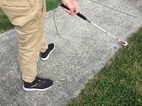 A close up image of a pair of male legs wearing beige coloured trousers, black runners and extending a white cane forward and to the right of the footpath against the edge of grass.
