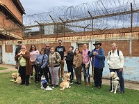 A group photograph of 12 people standing next to each other on grass, with a portion of the brick gaol wall seen in the background, including rusty spiralling barbed wire fencing extending beyond the brick fence above and behind our head height. Four dog guides and one Assistance Dog are also seen in close proximity to their handlers.