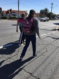 Frances is seen side profile and from behind crossing a light controlled street crossing with long cane and support cane