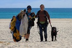 Garth being guided by his skydiving instructor on the sand with his dog guide Roy to his left and brilliant blue ocean in the background, the Parachuting gear is visible in the image